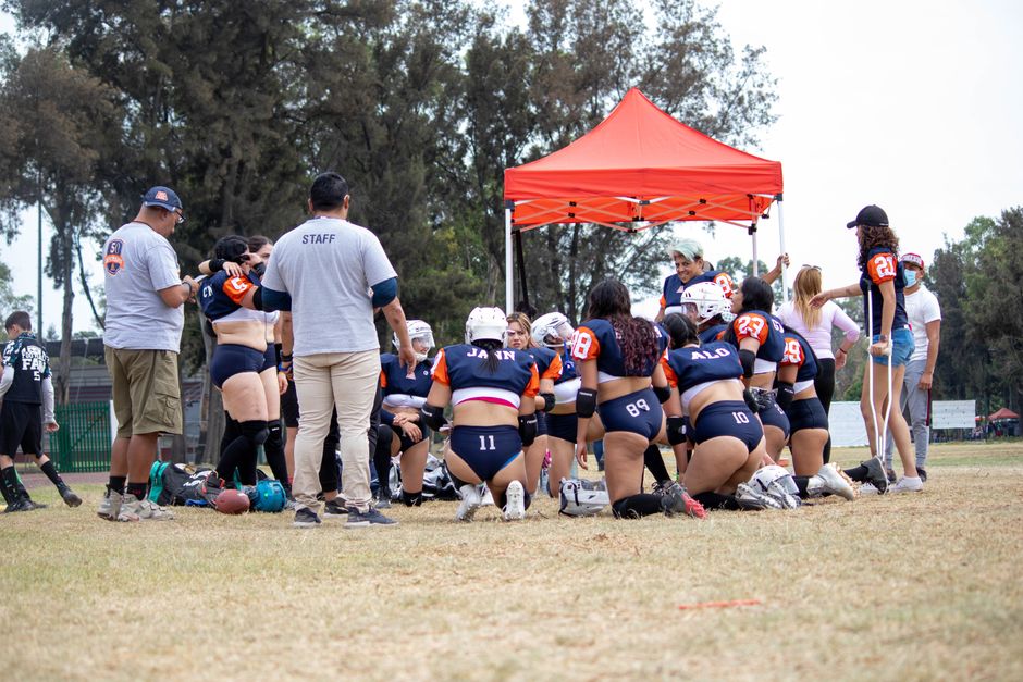 A women's flag football team huddles under a red canopy on a field, preparing for the game.